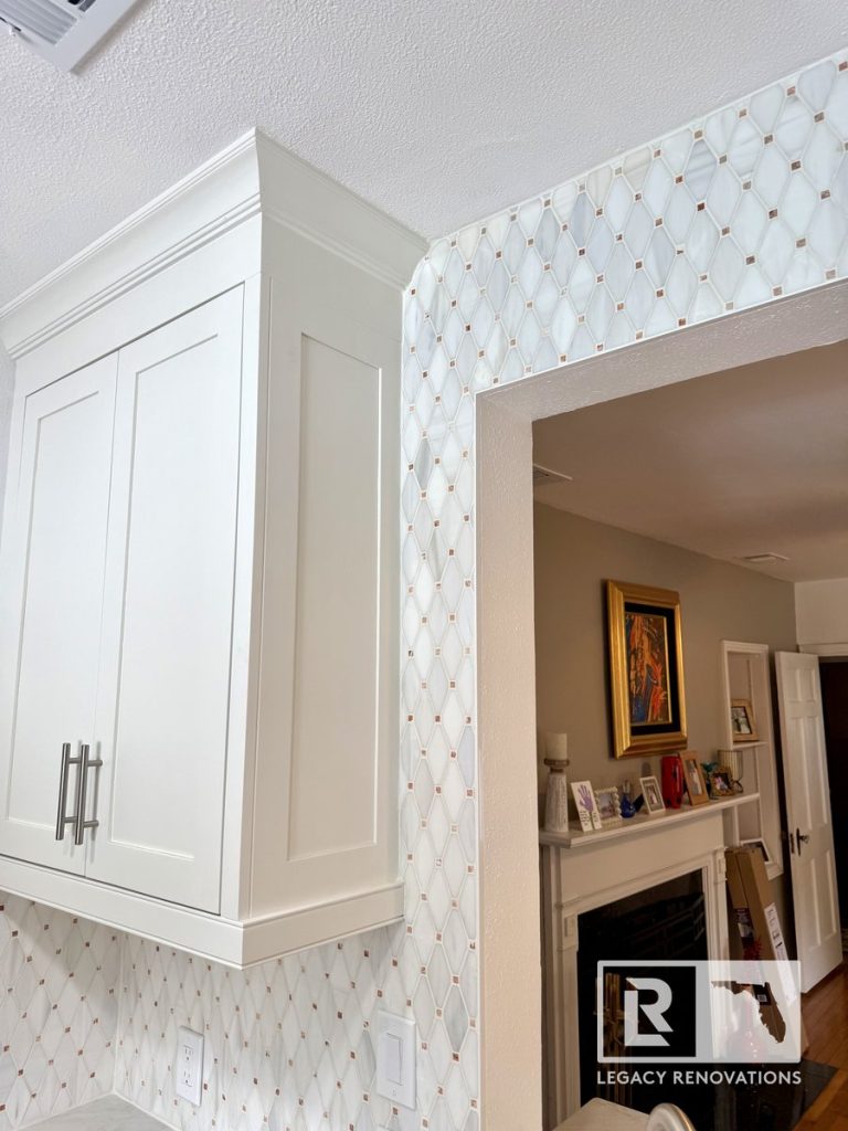 Kitchen remodel in Jacksonville, FL featuring tall white shaker-style cabinets, crown molding, a geometric tile backsplash, and a pass-through opening connecting the kitchen to the living area.