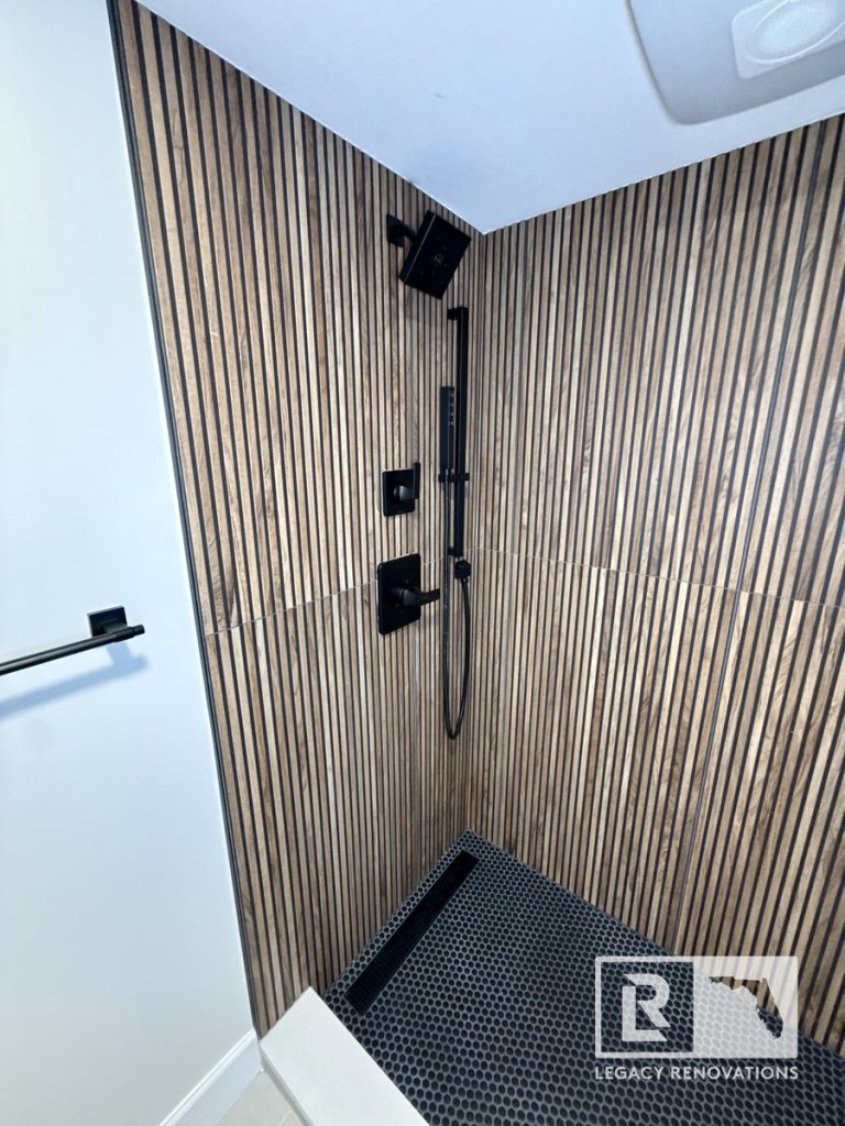 Walk-in shower with fluted wood-look tile walls, matte black rainfall shower system, linear drain, and black penny tile floor in Jacksonville, FL.