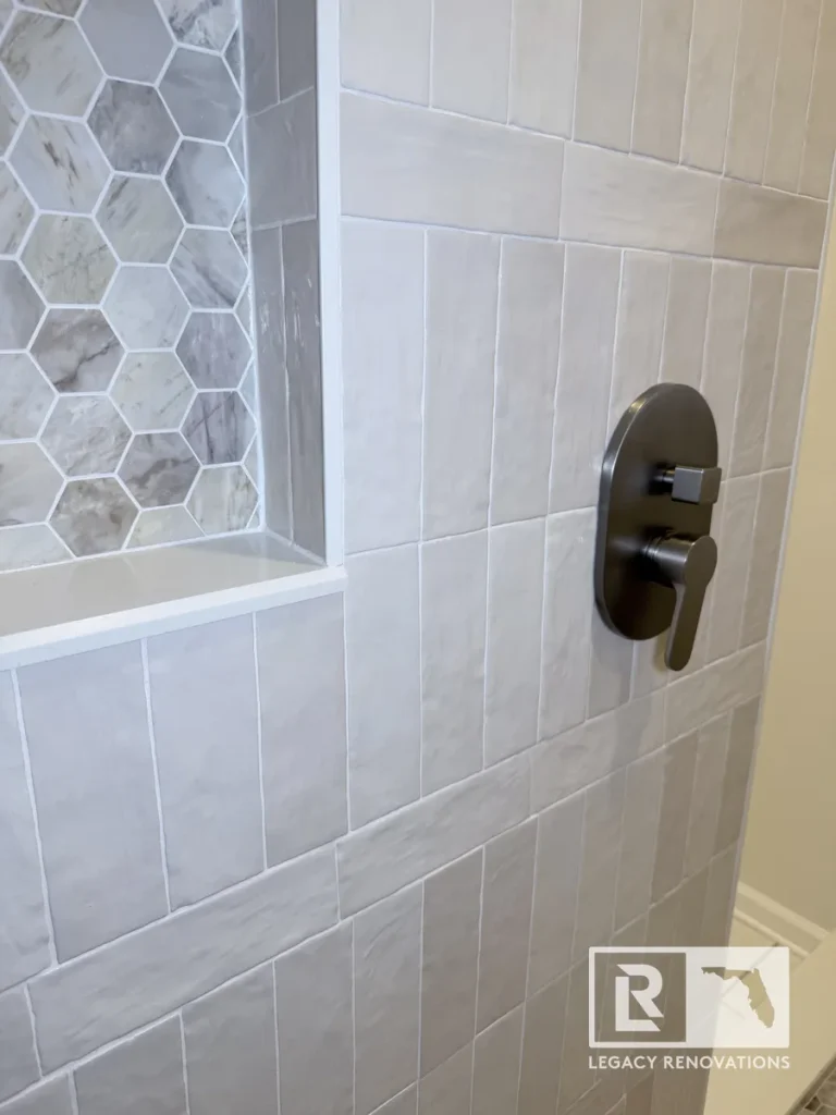 Oval dark metallic shower faucet handle plate mounted next to a marble hexagon tiled niche in a Mandarin Jacksonville FL residence.