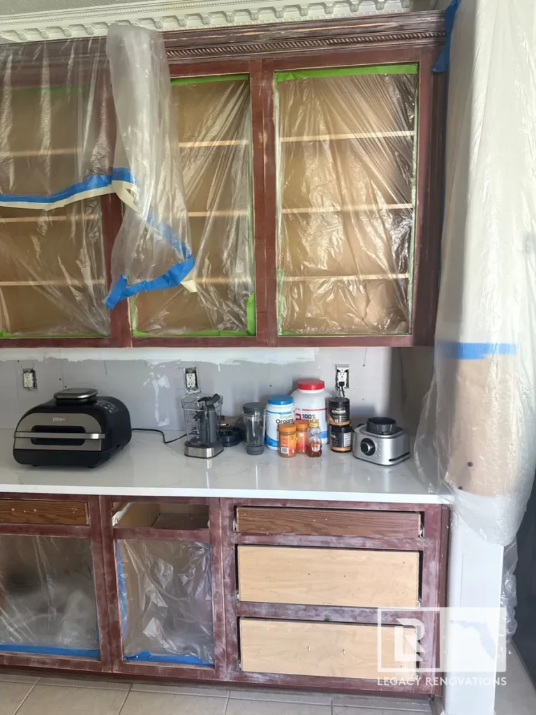 Sanded wooden kitchen cabinets taped off with clear plastic sheeting during refinishing.